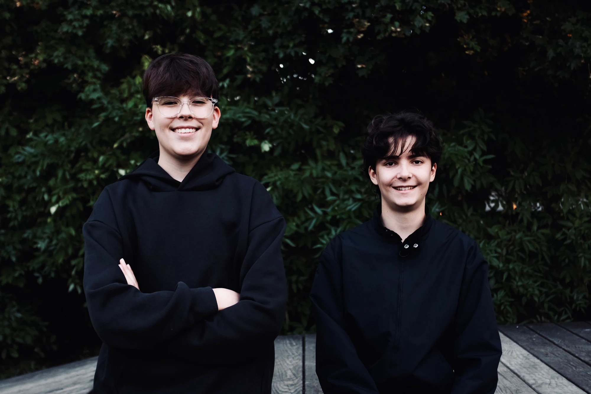 Aidan and Laurent outdoors in front of dark green foliage, both dressed in black and smiling.
